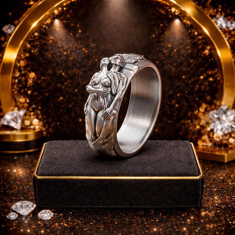 Peut inclure: Une bague argent&eacute;e avec un motif sculpt&eacute; de deux figures enlac&eacute;es. La bague a un large anneau et des d&eacute;tails en relief, mettant en valeur les formes des figures. La bague est pr&eacute;sent&eacute;e sur une surface en velours noir avec une bordure dor&eacute;e.