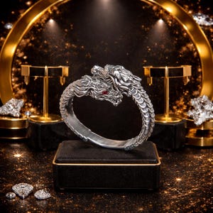 Peut inclure: Un bracelet argent&eacute; en forme de dragon avec des yeux en pierres pr&eacute;cieuses rouges. Le bracelet pr&eacute;sente deux t&ecirc;tes de dragon face &agrave; face, avec des d&eacute;tails d'&eacute;cailles complexes. Le bijou est expos&eacute; sur une bo&icirc;te noire avec une bordure dor&eacute;e.