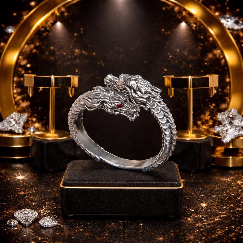 Peut inclure: Un bracelet argent&eacute; en forme de dragon avec des yeux en pierres pr&eacute;cieuses rouges. Le bracelet pr&eacute;sente deux t&ecirc;tes de dragon face &agrave; face, avec des d&eacute;tails d'&eacute;cailles complexes. Le bijou est expos&eacute; sur une bo&icirc;te noire avec une bordure dor&eacute;e.