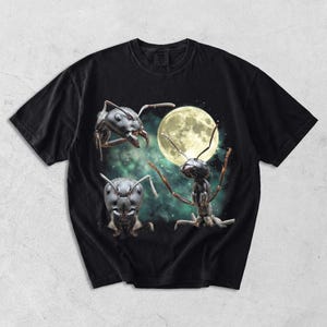 May include: Black t-shirt with a surreal graphic of three ants and a full moon. The ants are detailed with realistic textures and poses, set against a starry, green-tinged night sky. The shirt has a classic crew neck.