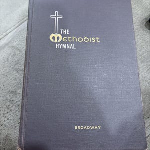 May include: A dark gray hardcover book titled The Methodist Hymnal with a gold cross symbol and the word Broadway printed in gold near the bottom. The cover has a textured fabric finish and the title text is centered in the upper portion.