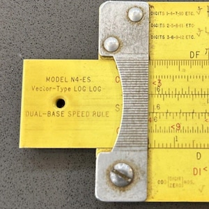 May include: A vintage yellow slide rule with a metal frame. The slide rule has black markings and text, including "MODEL N4-ES" and "DUAL-BASE SPEED RULE." The metal frame is silver and has rivets. The slide rule is a Vector-Type LOG LOG.