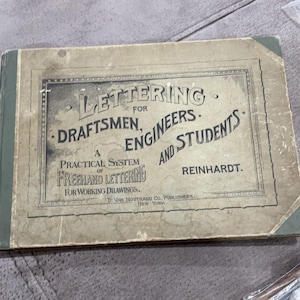 May include: A rectangular book with a weathered tan cover and a dark green spine. The front cover features black text reading: Lettering for Draftsmen, Engineers and Students, A Practical System of Freehand Lettering for Working Drawings, Reinhardt, D. Van Nostrand Co., Publishers, New York.