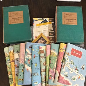 May include: Two green storage boxes labeled American Geographical Society Around the World Program, accompanied by a collection of illustrated travel booklets featuring maps and cultural scenes, plus a small portrait of John F. Kennedy.