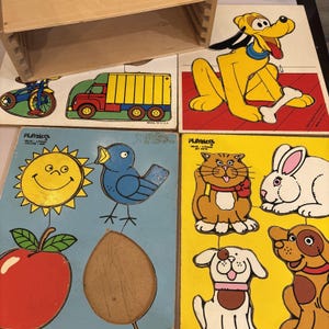 May include: A vintage Playskool wooden puzzle set featuring colorful animal and vehicle designs. The set includes a yellow dog with a bone, a red truck, a blue bird, a smiling sun, and other shapes. The puzzle pieces are displayed with a wooden storage box.