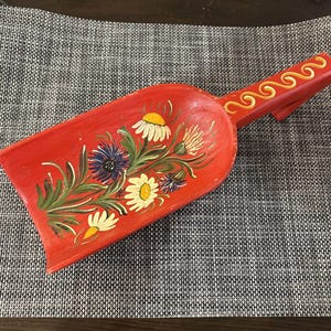 May include: A red wooden scoop featuring a hand-painted floral design with white daisies, blue cornflowers, and green leaves. The handle is decorated with a yellow wavy line pattern. The scoop has a wide, flat base and a long, sturdy handle.