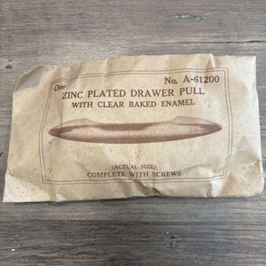 May include: A vintage zinc-plated drawer pull with clear baked enamel, packaged in a brown paper wrapper. The package includes the text "ZINC PLATED DRAWER PULL" and "COMPLETE WITH SCREWS".