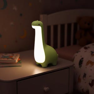 Può includere: Una lampada da notte verde a forma di dinosauro con un lungo collo e una luce bianca brillante. La lampada è su un comodino bianco in una stanza poco illuminata. Un'aggiunta divertente e funzionale per la cameretta.
