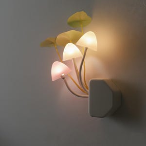 May include: A mushroom-shaped night light with three illuminated mushroom caps in pink and white. The light has a white base and stems, with green leaf accents. It plugs directly into a wall socket.
