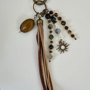 May include: A keychain with a bronze-colored ring and various charms. The charms include a brown oval stone, a leather tassel, beaded strands with green, brown, and white beads, and a sun-shaped charm with a white center.