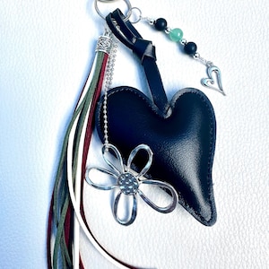 May include: A keychain featuring a dark blue leather heart, a silver-toned flower charm, and a multi-colored tassel with olive green, white, and maroon strips. A silver chain holds black and light green beads and a small silver heart-shaped charm.