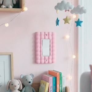 Könnte beinhalten: Ein rosa Lichtschalter mit einer strukturierten Abdeckung und einem weißen Schalter. Die Abdeckung ist rechteckig und an einer rosa Wand befestigt. Ein dekoratives Mobile und Kinderbücher sind im Hintergrund zu sehen.