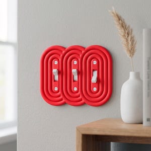 Könnte beinhalten: Drei rote Lichtschalterabdeckungen mit weißen Schaltern sind an einer hellgrauen Wand montiert. Die Abdeckungen haben ein ovales, geschichtetes Design. Eine weiße Vase mit einer getrockneten Pflanze und ein Holztisch befinden sich im Hintergrund.