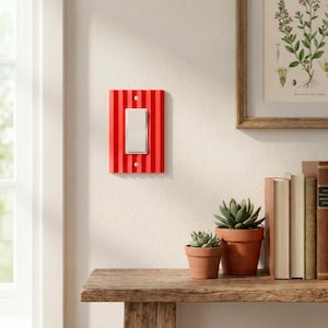 May include: A red and white striped light switch cover with a silver switch. The cover is mounted on a white wall. The image also shows a wooden shelf with potted plants and books.