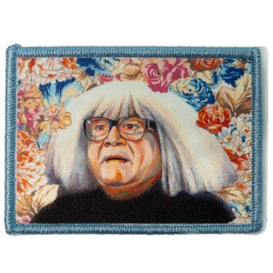 May include: Embroidered patch featuring a portrait of a person with white hair and glasses, set against a floral background of pink, blue, and orange flowers. The patch has a blue border.