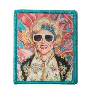 May include: Embroidered patch featuring a portrait of a woman wearing sunglasses, a floral jacket, and a jeweled headband. The background is a colorful floral pattern with a teal border. The patch is rectangular in shape.