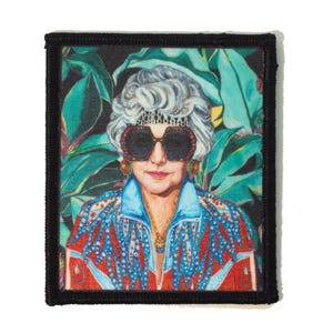 May include: A square patch with a black border features a portrait of a woman with sunglasses and a patterned jacket. The background includes green tropical leaves. The woman has gray hair and is wearing a necklace and earrings. The patch is likely for clothing or accessories.