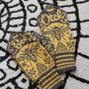 May include: A pair of grey and yellow knitted mittens with a bee and honeycomb pattern. The mittens have a striped cuff.