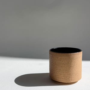 May include: A cylindrical ceramic cup featuring a textured, light tan exterior surface and a smooth, glossy black interior finish. The vessel has a straight-sided profile with a flat base and a simple, unadorned rim, showcasing a contrast in color.