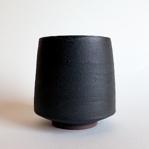 May include: A handmade, black ceramic vase with a textured surface. The vase has a wide, cylindrical shape that tapers slightly towards the base, which rests on a small, reddish-brown ring. The background is a plain white.