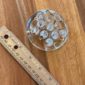 May include: A clear glass flower frog with multiple circular openings for stems, positioned next to a wooden ruler showing a diameter of approximately 3 inches. The circular glass piece features a smooth, transparent surface with distinct indentations.