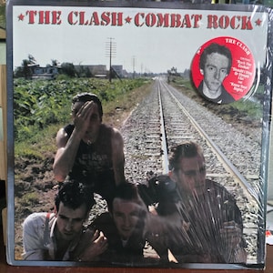 May include: The Clash's "Combat Rock" album cover. The image features the band members posing on a railway track. The album title is in red and white text. A circular sticker with a band member's portrait is on the right.