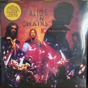 Può includere: Copertina dell'album "MTV Unplugged" degli Alice in Chains. La copertina dell'album presenta i membri della band e il testo "Alice in Chains" e "Unplugged". La copertina include anche il testo "The Complete Show" e i titoli delle canzoni.