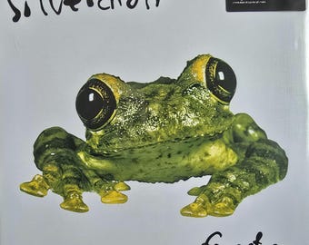 Puede incluir: Una portada de disco de vinilo con una rana verde de grandes ojos negros centrada sobre un fondo blanco. El texto silverchair está impreso en negro en la parte superior, y frogstomp está impreso en negro en la parte inferior derecha. Una pegatina negra dice Music On Vinyl.