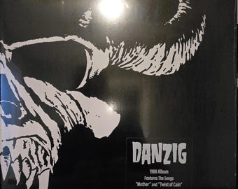 May include: A black and white vinyl record cover featuring a stylized skull graphic on the left side. The text reads DANZIG, 1988 Album, Features The Songs Mother and Twist of Cain. The square cover is encased in a clear plastic protective sleeve.
