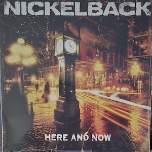 Może przedstawiać: Okładka płyty winylowej z napisem NICKELBACK u góry i HERE AND NOW u dołu. Grafika przedstawia ciemną, nocną scenę ulicy miejskiej z wyraźną wieżą zegarową, świecącymi latarniami ulicznymi i smugami światła na jezdni.
