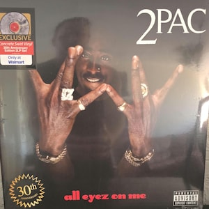 May include: A 2Pac All Eyez On Me 30th Anniversary Edition 2LP vinyl record set with a concrete swirl vinyl design. The cover features a portrait of 2Pac making a hand gesture, with the text 2PAC, all eyez on me, and a Parental Advisory label.