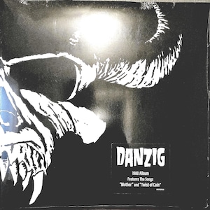 May include: A black and white vinyl record cover featuring a stylized skull graphic. The text reads: DANZIG, 1988 Album, Features The Songs, Mother and Twist of Cain. The artwork is high-contrast with a dark background and white illustrative details.