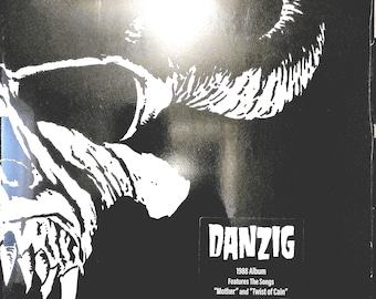 Puede incluir: Una cubierta de disco de vinilo en blanco y negro con un gráfico de calavera estilizado. El texto dice: DANZIG, 1988 Album, Features The Songs, Mother and Twist of Cain. La obra es de alto contraste con un fondo oscuro y detalles blancos.