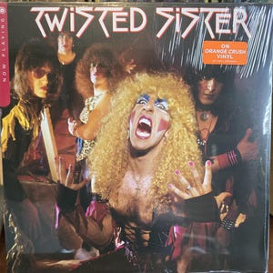 Puede incluir: Un álbum de vinilo de Twisted Sister, con los miembros de la banda en una foto de grupo. La portada del álbum tiene el nombre de la banda en una fuente blanca estilizada. El álbum está sellado en plástico.
