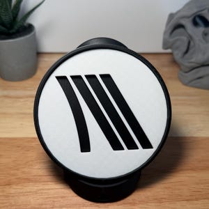 May include: A black and white round phone grip with a black border. The white center features four black, angled lines. The grip is on a wooden surface, with a small potted plant and a gray sculpture in the background.