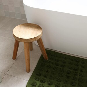 May include: A small, round wooden stool with three legs stands next to a green bath mat with a textured, floral pattern. The stool is made of light-coloured wood and is positioned near a white bathtub in a bathroom setting.