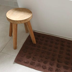 May include: A small, round wooden stool with three legs stands beside a brown bath mat with a textured, floral pattern. The stool is made of light-coloured wood and the mat is rectangular.