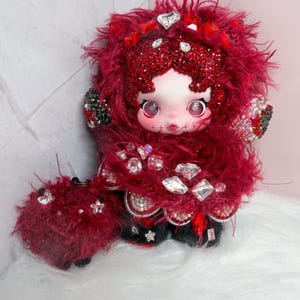 May include: A handmade doll with a pale face, large eyes, and stitched details. The doll is adorned with red fluffy material, sparkling gems, and black accents. The doll's outfit is embellished with rhinestones and red accents.