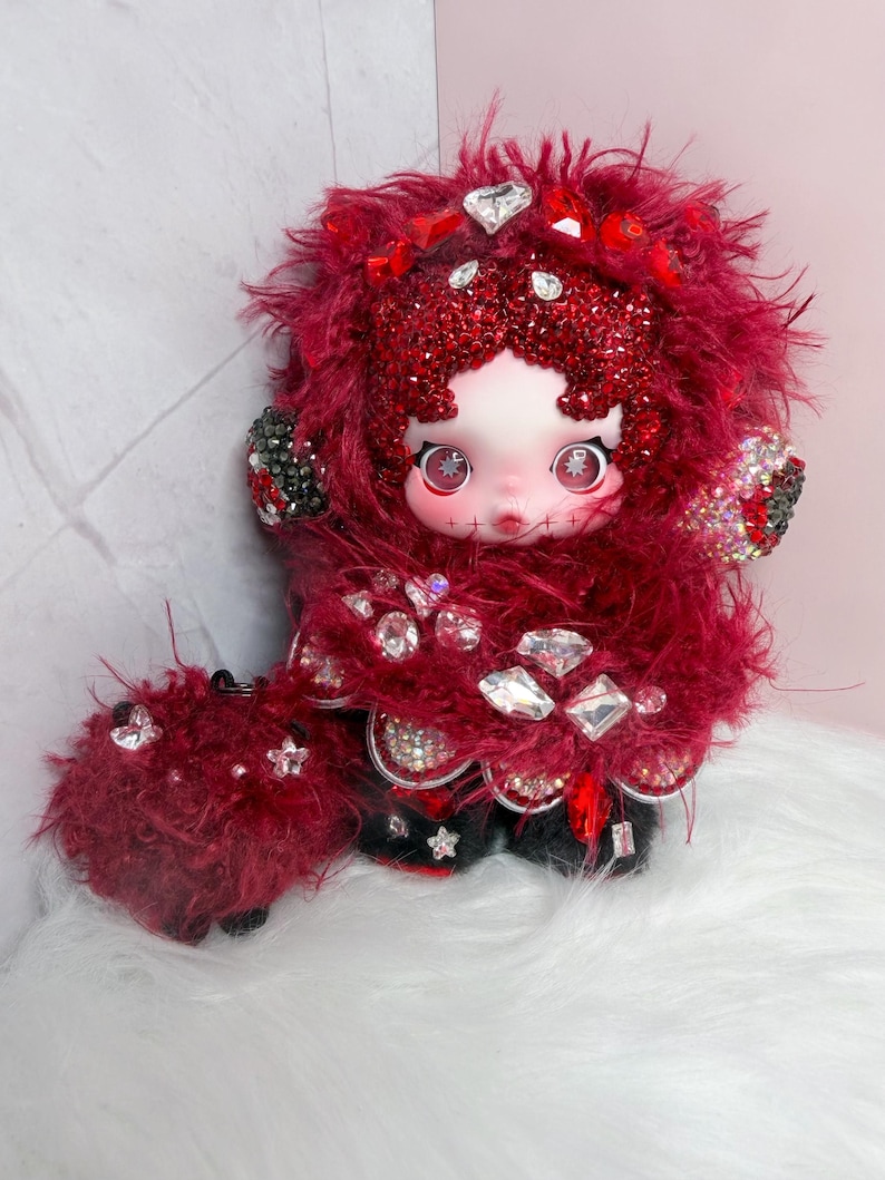 May include: A handmade doll with a pale face, large eyes, and stitched details. The doll is adorned with red fluffy material, sparkling gems, and black accents. The doll's outfit is embellished with rhinestones and red accents.