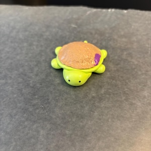 Puede incluir: Figura de tortuga hecha a mano con cuerpo verde claro, caparazón marrón texturizado y un pequeño acento morado. La tortuga tiene una cara sonriente con ojos de puntos negros. La figura probablemente está hecha de arcilla o polímero.