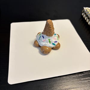 May include: A whimsical, handmade clay turtle figurine. The turtle has a brown shell and limbs, with a white frosting-like topping adorned with colorful sprinkles. A brown ice cream cone sits atop the turtle's back, completing the dessert-themed design.