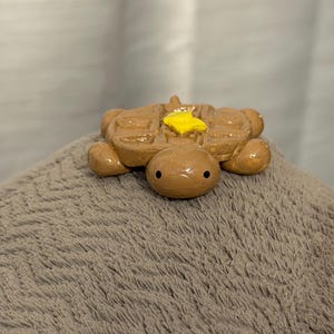 Puede incluir: Una caprichosa figura de tortuga marrón con un caparazón de gofre y una mota de mantequilla amarilla en la espalda. La tortuga tiene pequeños ojos negros y descansa sobre una superficie gris texturizada. Un artículo decorativo único.