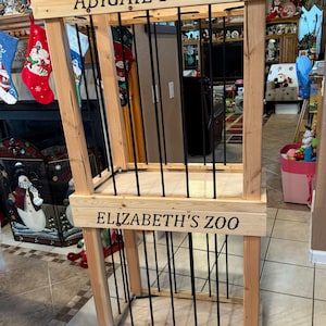 May include: A wooden zoo enclosure with the text "ABIGAIL'S ZOO" and "ELIZABETH'S ZOO" in black lettering. The structure features vertical black bars and a natural wood frame. The enclosure is designed for decorative purposes.