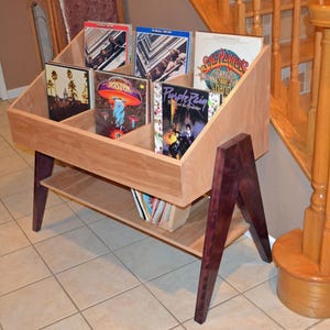 Può includere: Un mobile porta dischi in legno con finitura marrone chiaro, con scomparti angolati per dischi in vinile. L'unità ha gambe viola scuro e un ripiano inferiore, che mostra diverse copertine di album. Il design è moderno.