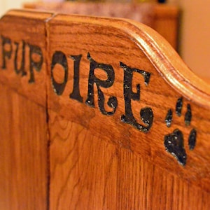 May include: Close-up of a wooden pet bed with the word "PUPOIRE" in black lettering. A black paw print is also visible. The bed is made of light brown wood with a curved top.