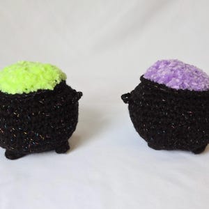 May include: Two crocheted cauldrons made of black yarn with subtle iridescent flecks. One cauldron features a bright neon green textured top, while the other features a soft lavender textured top. Both items have small side handles and three feet.