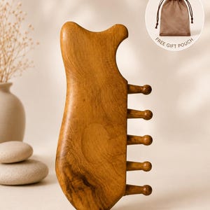 May include: A wooden gua sha tool with a unique shape, featuring a curved top and five rounded prongs. The tool is a warm brown color and appears to be made of natural wood. A small brown gift pouch is included. The text "FREE GIFT POUCH" is also visible.