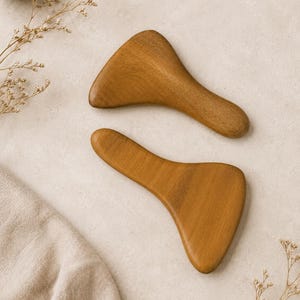 May include: Two wooden gua sha tools with a smooth, curved design. The tools are a warm brown color and are positioned on a neutral-toned surface. Dried sprigs and a small bowl are also visible.