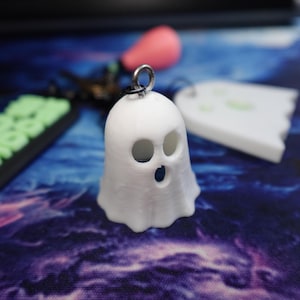 May include: A small, white, three-dimensional ghost figurine featuring two circular eye cutouts and one circular mouth cutout. The ghost has a smooth, rounded top and a scalloped bottom edge, with a silver-toned metal jump ring attached to the top.