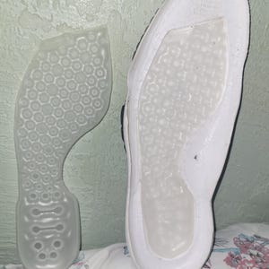 May include: A white shoe insole with a translucent, semi-rigid plastic insert featuring a repeating hexagonal honeycomb pattern. The insert is shown both detached and placed within the white foam base of the shoe, which has text printed at the heel.